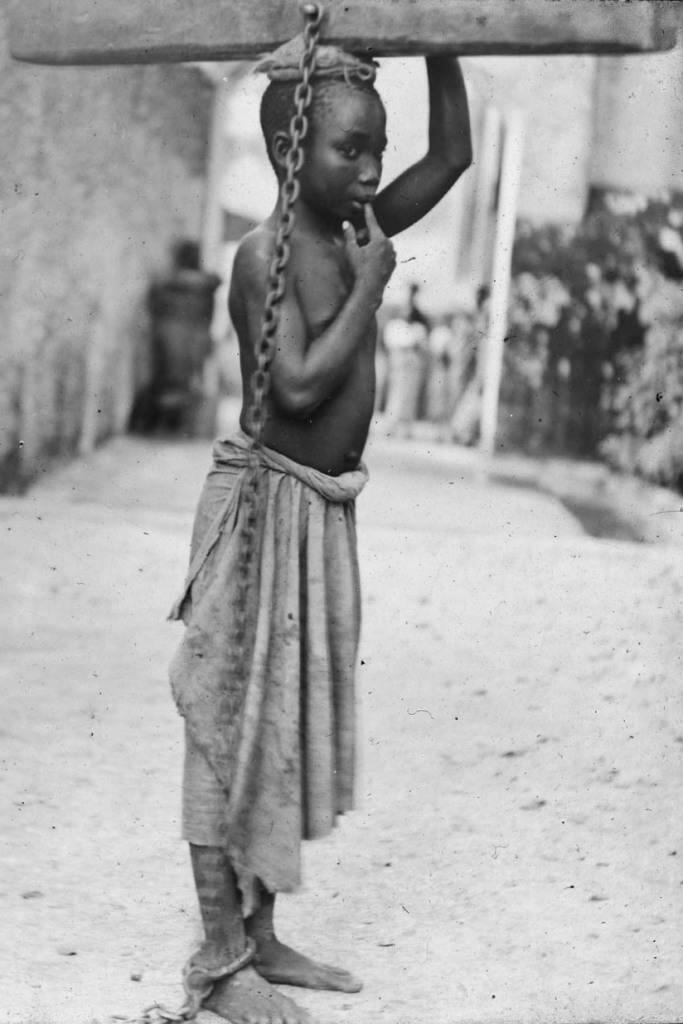 1890. Zanzibári rabszolga fiú. Arab gazdája egy kisebb kihágás miatt bűntette a rönkhöz láncolással. Ezt a fején hordozva a legkönnyebb mozognia..jpg